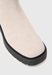 Beige semsket sko med avrundet tå og tykk svart gummisåle med mønster, vist fra en litt skrå nærbilde.