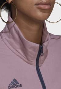 Lichtpaarse jas met ritssluiting, gemaakt van een gestructureerde stof, hoge kraag en een marineblauwe rits. Voorzien van een geborduurd zwart Adidas-logo.