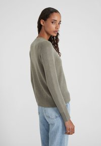 Jeune femme aux cheveux bouclés portant un pull en maille vert olive et un jean bleu clair, se tenant dans une pose de trois-quarts de dos.