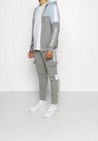 Grauer und hellblauer Kapuzenpullover mit Reißverschluss und Text auf den Ärmeln, kombiniert mit passenden grauen Jogginghosen mit Cargotaschen und weißen Akzenten. Weiße Turnschuhe.