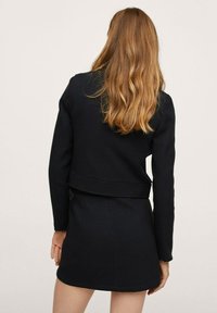 Chaqueta negra ajustada con mangas largas, que presenta una cintura entallada y una textura suave, combinada con una falda corta a juego.