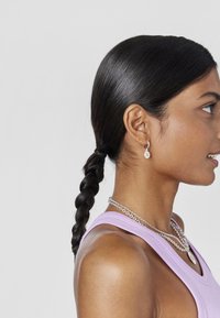 Perfil lateral de una persona con pelo largo y oscuro trenzado, que lleva una camiseta sin mangas de color lilac, collares de plata en capas y pequeños aretes colgantes.