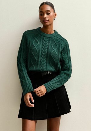 Pull en maille torsadée vert foncé avec un col rond et des poignets côtelés, associé à une jupe plissée noire et à une ceinture en accent.