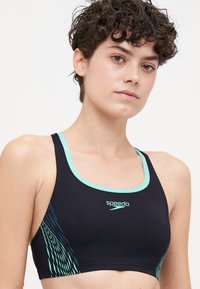 Sportovní podprsenka Speedo v černé barvě s tyrkysovými akcenty. Má design s překříženými ramínky, hladký materiál a grafický vzor na straně.