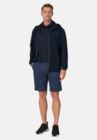 Mann steht vor neutralem Hintergrund, trägt eine marineblaue Jacke mit Reißverschluss, ein marineblaues Poloshirt, blaue Shorts und schwarze Sneakers.