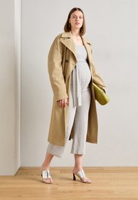 En tan trenchcoat over en grå jumpsuit med midjebånd, kombinert med sølvhælede sandaler og en grønn clutch, på et tregulv.