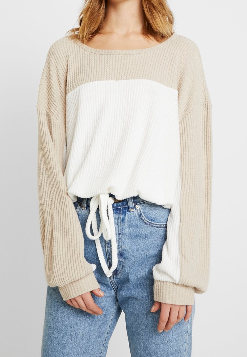 Pull à côtes beige et blanc avec une coupe décontractée, manches larges et cordon de serrage ajustable à la taille. Un jean en denim complète le look.