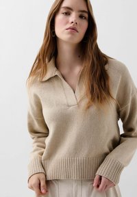 Beige gebreide trui met een kraag en een gestreepte zoom. Lange mouwen met een ontspannen pasvorm en subtiele textuurdetails.