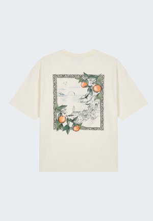 Camiseta color crema con un boceto enmarcado de un pueblo costero, con ramas de frutas naranjas y hojas verdes superpuestas en las esquinas superior e inferior.