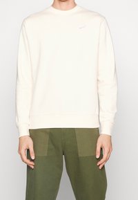 Světle broskvová mikina s dlouhými rukávy, žebrovanými manžetami a lemem, s malým bílým logem Nike na hrudi.