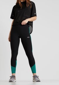 Czarna siateczkowa koszulka sportowa z krótkim rękawem, zestawiona z czarnymi legginsami z akcentami w kolorze teal. Szare buty sportowe dopełniają strój.