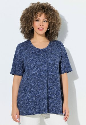 Mujer con cabello rizado que lleva una camiseta azul con estampado de manga corta y pantalones blancos, de pie frente a un fondo blanco.