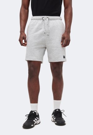 Άνδρας που φοράει ανοιχτό γκρι σορτς με κορδόνι, μαύρο μπλουζάκι, λευκές κάλτσες και μαύρα αθλητικά παπούτσια Adidas, στέκεται μπροστά σε ένα απλό φόντο.