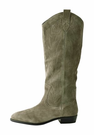 Personne portant une veste en daim marron, une robe noire et des bottes en daim vert clair montant jusqu'aux genoux, marchant sur un trottoir pavé près de murs en briques et en pierre.