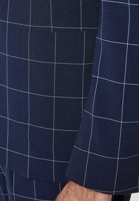 Navy blue suit jacket with white grid pattern, featuring a patch pocket and textured fabric. Tailored fit with clean lines and minimal accents.