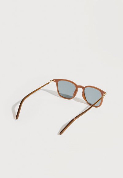 Καφέ γυαλιά ηλίου με τετράγωνα γκρι φακούς και λεπτά πλαίσια, τοποθετημένα σε απλή λευκή επιφάνεια.