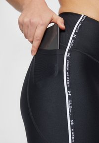 Leggings sportivi neri con una tasca laterale che ospita uno smartphone. Presentano un logo del marchio bianco e un accento lungo la cucitura laterale. Texture morbida.