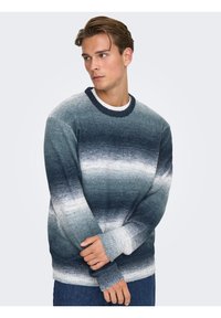 Jeune homme portant un pull à dégradé bleu et blanc, regardant vers sa gauche, les bras croisés devant lui, sur un fond clair uni.