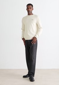 Hombre de pie con un suéter largo de manga crema, pantalones gris oscuro y zapatos negros contra un fondo blanco plano.