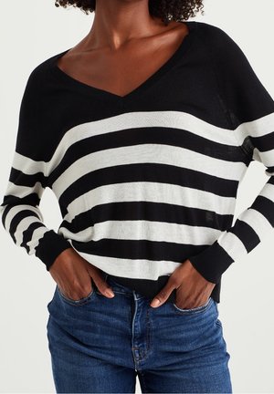 Persona vistiendo un suéter de rayas horizontales en blanco y negro con cuello en V y jeans azules, con las manos en los bolsillos frente a un fondo liso.