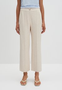 Pantalones beige de pierna ancha con tejido texturizado, que presentan plisados frontales y un cierre con botón. Llevados con sandalias planas y sencillas.