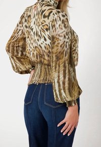 Blouse imprimée avec un motif animalier doré et marron, tissu transparent, manches longues bouffantes, taille smockée ; associée à un jean en denim bleu foncé.
