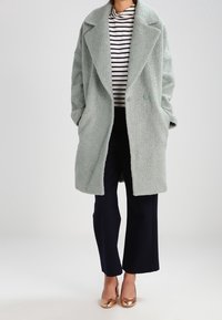 Cappotto testurizzato blu chiaro con ampio colletto e doppi bottoni, indossato sopra un dolcevita a righe e pantaloni ampi blu navy.