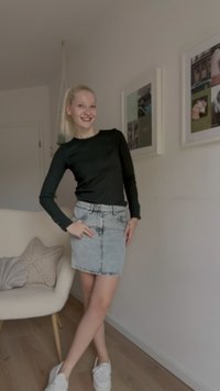 Blusa preta de manga longa com design ajustado, combinada com uma saia mini de ganga clara com fecho de botão e zíper, textura suave, e sapatilhas casuais.