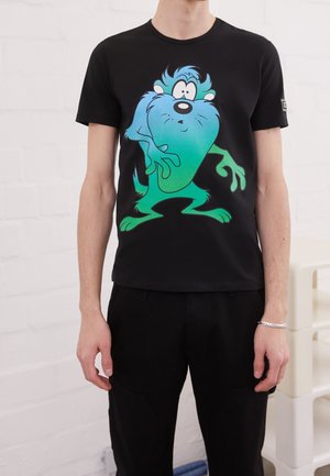Personne portant un t-shirt noir avec un grand graphique de personnage de dessin animé bleu et vert sur le devant, debout à l'intérieur.