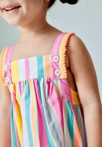 Monivärinen raidallinen mekko, jossa on vaaleanpunaista, keltaista, turkoosia ja valkoista. Koristeltu olkaimet, joissa on pom-pom-reunus ja napit.