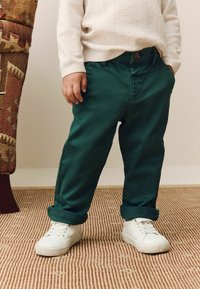 Un enfant portant un pantalon vert retroussé, un pull blanc et des baskets blanches se tient sur un tapis texturé à côté d'une jambe de chaise à motifs.