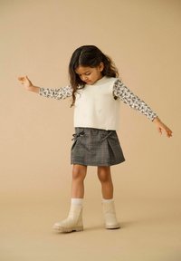 Młoda dziewczynka balansująca z wyciągniętymi rękami, ubrana w biały bezrękawnik, wzorzystą bluzkę z długim rękawem, spódniczkę w kratę i beżowe buty.