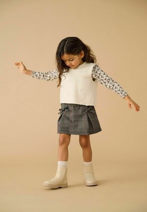 Meisje dat balanceert met uitgestrekte armen, gekleed in een witte mouwloze sweater, een gestreepte langemouwen shirt, een geruite rok en beige laarzen.