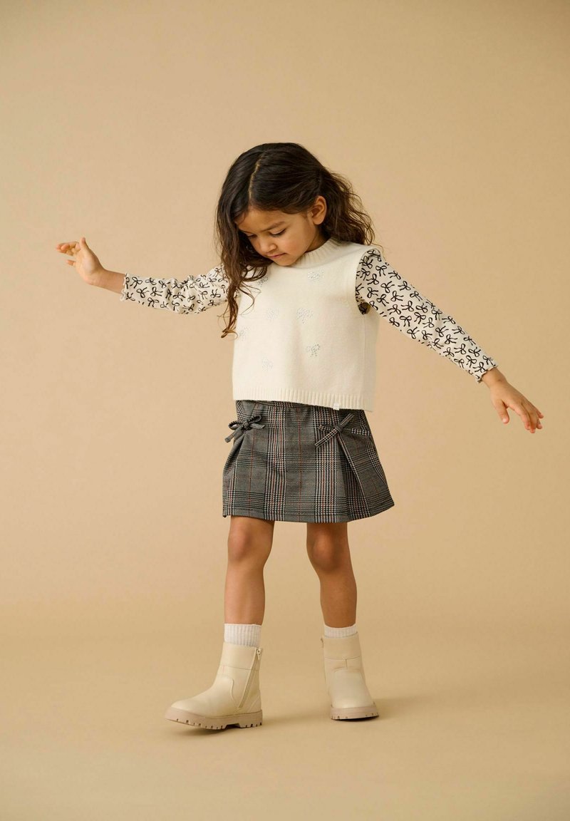 Młoda dziewczynka balansująca z wyciągniętymi rękami, ubrana w biały bezrękawnik, wzorzystą bluzkę z długim rękawem, spódniczkę w kratę i beżowe buty.