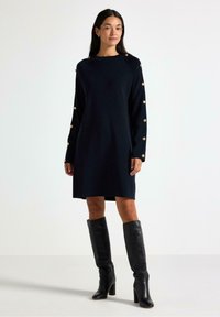 Abito navy con maniche lunghe, caratterizzato da bottoni dorati lungo le maniche. L'abito ha una vestibilità rilassata e un colletto rotondo. Abbinato a stivali neri al ginocchio.