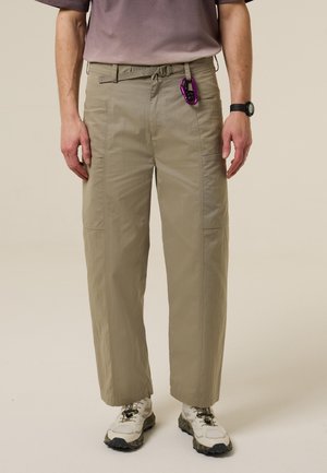 Person trägt beige Cargohosen mit einem kleinen lila Karabiner am Gürtelschlaufen und weißen Wanderschuhen.