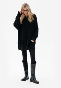 Overgrote zwarte gebreide trui met verlaagde schouders, geribde zoom, gecombineerd met zwarte leggings en glanzende zwarte laarzen tot de knie.