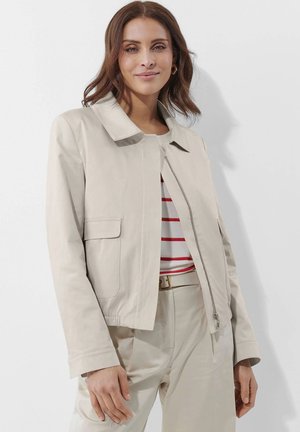 Light beige cropped jacket with a flat collar, side pockets, and a front zipper. Worn over a striped shirt and matching beige pants.