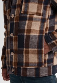 Rutet flanneljakke i brune, marine- og beige farver, med knaplukning, brystlomme og blød tekstur.
