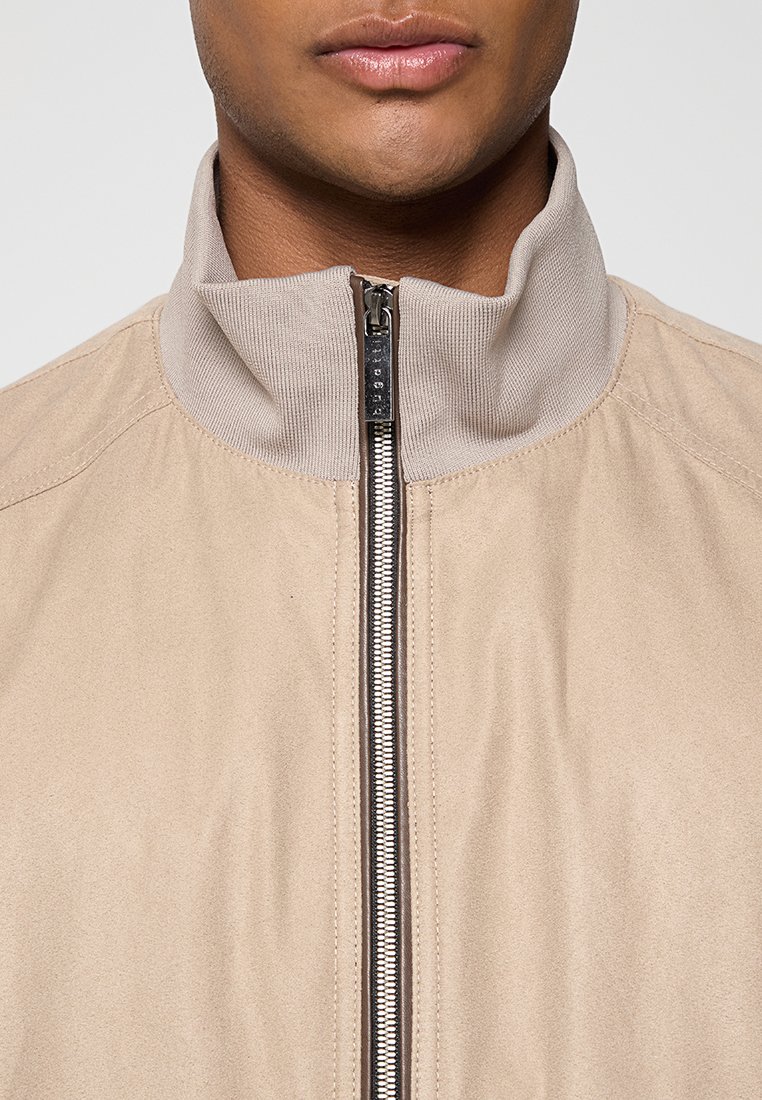 Veste bomber beige avec un col montant, en tissu texturé, et une fermeture éclair en métal argenté à l'avant. Présente des détails de couture sport.