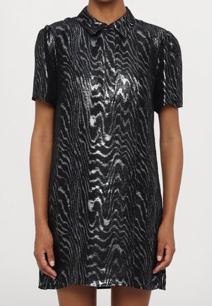 Sort kortærmet kjole med skinnende sølv bølgemønster og spids krave, båret af en person stående mod en ensfarvet baggrund.