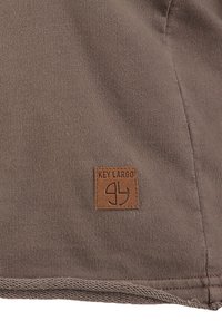 Pezzo di tessuto marrone con il testo in rilievo "KEY LARGO" e un logo. Il patch è cucito su una superficie di tessuto liscia e opaca.