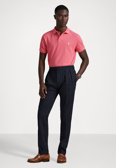 Camisa polo rosa com dois botões, calças listradas em azul-marinho e sapatos de couro castanho. A camisa apresenta um logo no lado esquerdo do peito.