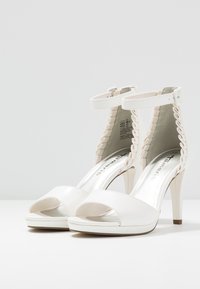Bílé sandály na vysokém podpatku s otevřenou špičkou, pásky kolem kotníku a dekorativním proplétaným detailem podél paty. Hladký materiál a lesklá podr�ážka.