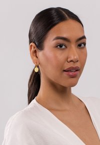 Goldene Creolen mit einem strukturierten runden Anhänger, der aufwendige Details aufweist, getragen von einer Person mit glattem Haar.