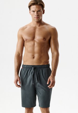 Mężczyzna bez koszulki o jasnobrązowych włosach, ubrany w ciemnoszare szorty z troczkami, stoi zwrócony przodem na jednolitym białym tle.