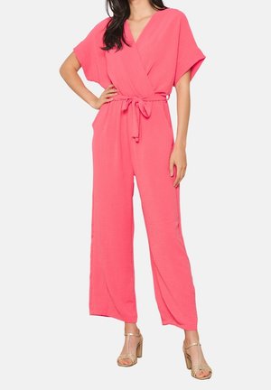 Vrouw in een felroze jumpsuit met korte mouwen en brede pijpen, met een ceintuur, en beige hoge hakken op een witte achtergrond.