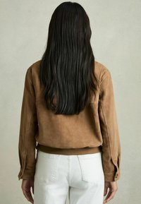Jachetă din suede maro cu o croială lejeră, cu mâneci lungi și un tiv care ajunge la talie. Asortată cu pantaloni albi.