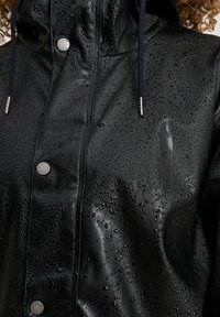 Musta vedenpitävä takki, jossa on kiiltävä ja sileä pinta, harmaita neppareita ja huppu tummilla nyöreillä, joiden pinnalla näkyy vesipisaroita.