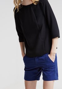 Osoba v černém tričku s tříčtvrtečními rukávy a modrých šortkách po kolena, se rukama v kapsách na jednobarevném pozadí.